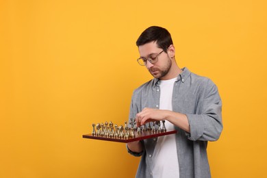 Handsome man holding chessboard with game pieces on orange background. Space for text