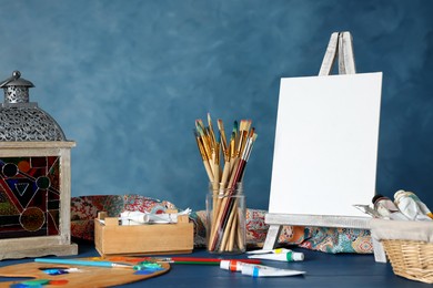 Photo of Many brushes, easel with blank canvas and paints on blue wooden table