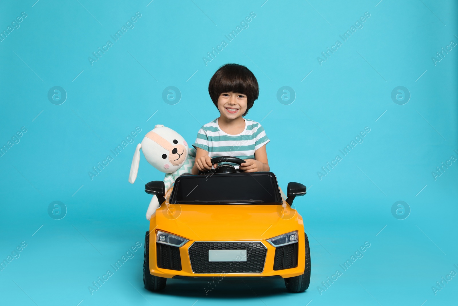 Photo of Cute little boy with toy bunny driving children's car on light blue background