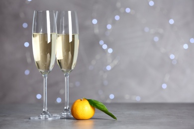 Sparkling wine in glasses near tangerine on grey table against blurred lights, space for text