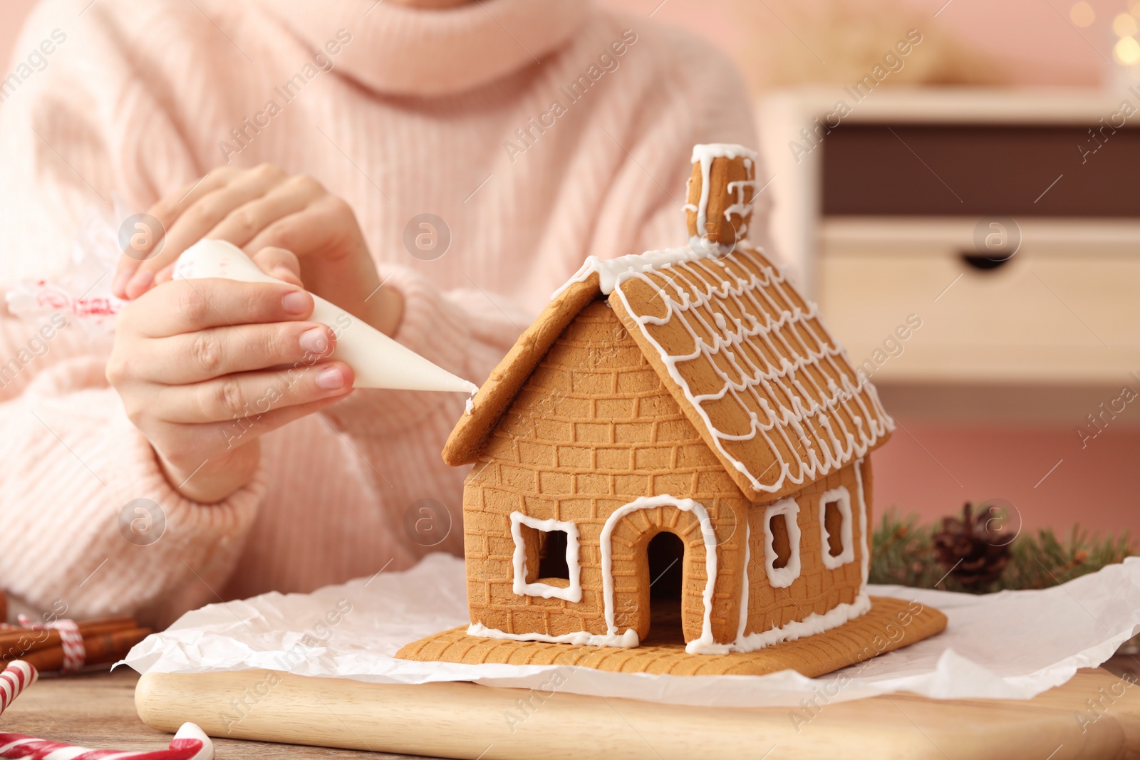 Photo of Woman decorating gingerbread house with icing at table, closeup