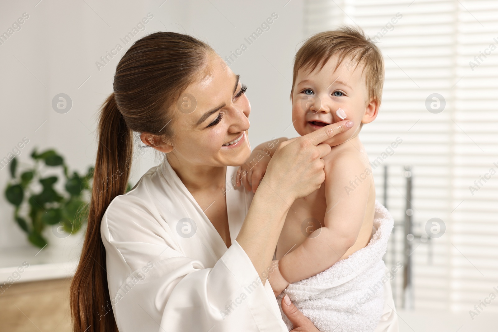 Photo of Happy mother applying moisturizing cream onto baby`s face in bathroom