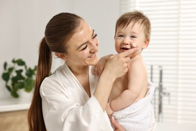 Happy mother applying moisturizing cream onto baby`s face in bathroom