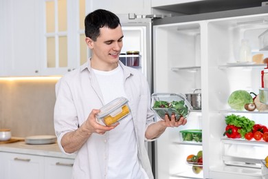 Happy man holding containers with vegetables near refrigerator in kitchen