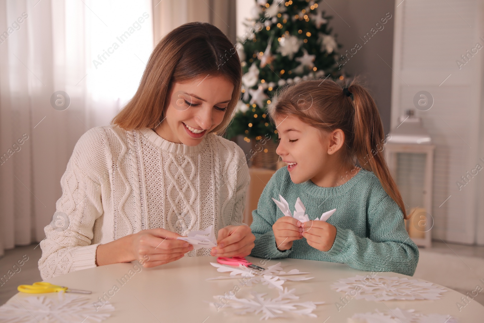 Photo of Happy mother and daughter making paper snowflakes at table near Christmas tree indoors