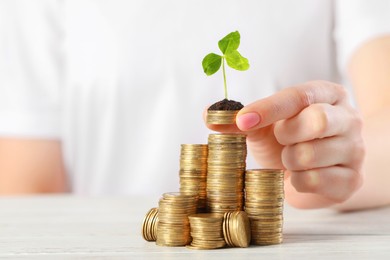 Photo of Woman putting coins with green sprout onto stack at white table, closeup. Investment concept