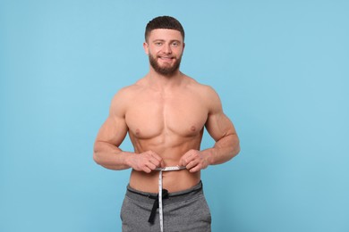 Photo of Portrait of happy athletic man measuring waist with tape on light blue background. Weight loss concept