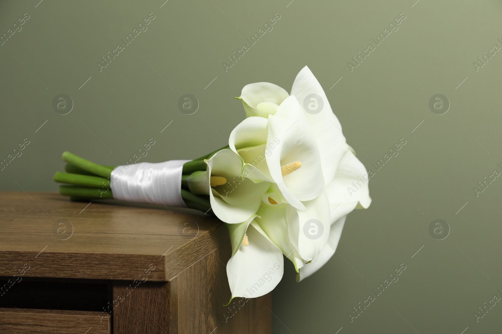 Photo of Beautiful calla lily flowers tied with ribbon on wooden table near olive wall
