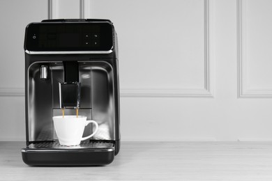 Photo of Modern espresso machine pouring coffee into cup on white wooden table near light wall. Space for text