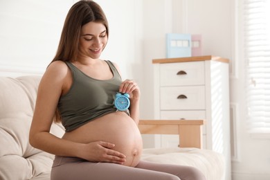 Photo of Young pregnant woman holding alarm clock near her belly at home, space for text. Time to give birth