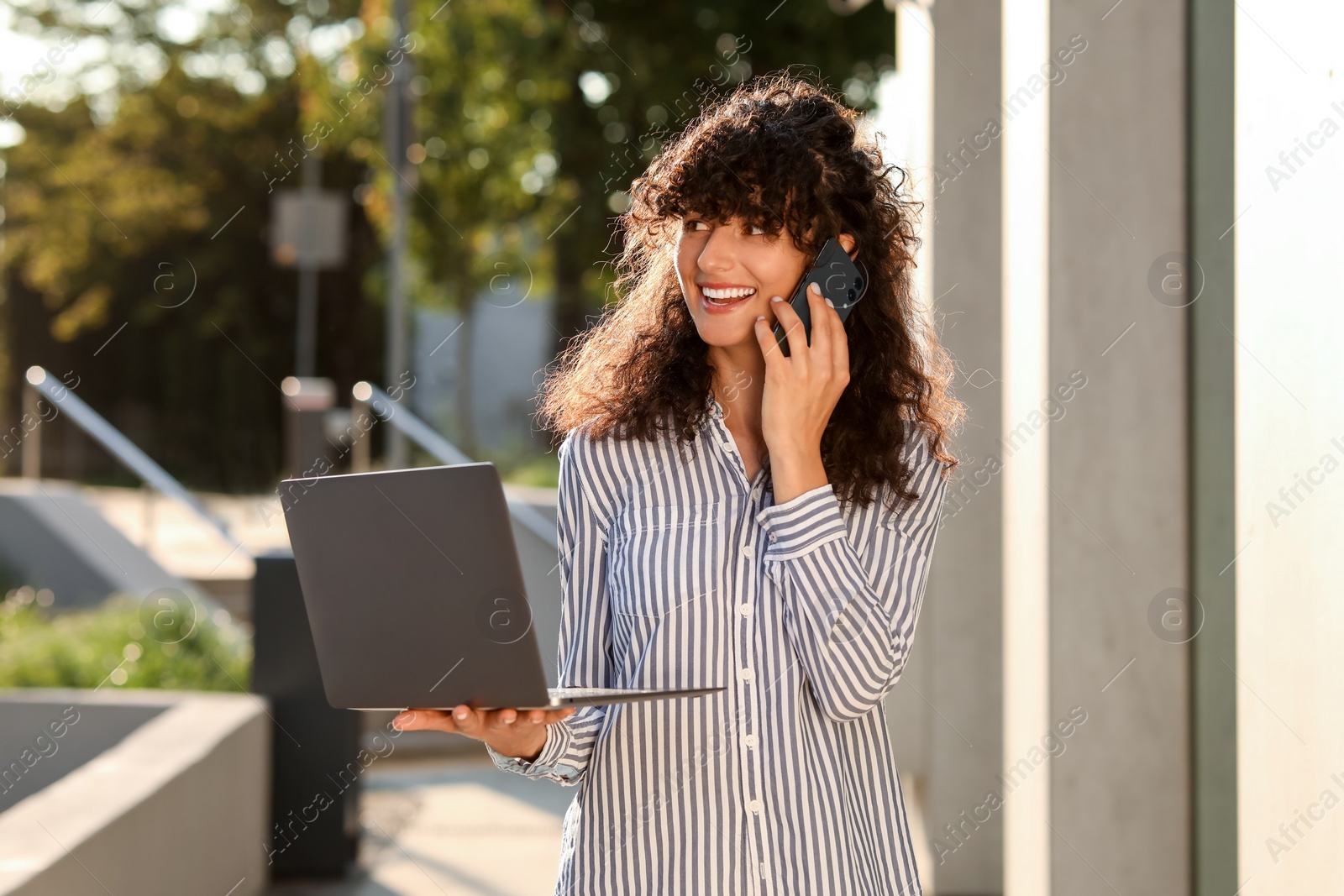 Photo of Happy young woman with modern laptop and talking on smartphone outdoors