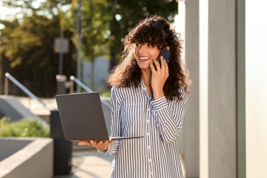 Happy young woman with modern laptop and talking on smartphone outdoors