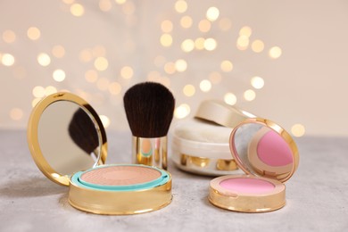 Photo of Face bronzer, blusher and other cosmetic products on grey textured table against blurred lights, closeup