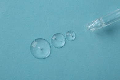 Dripping cosmetic serum from pipette onto light blue background, top view
