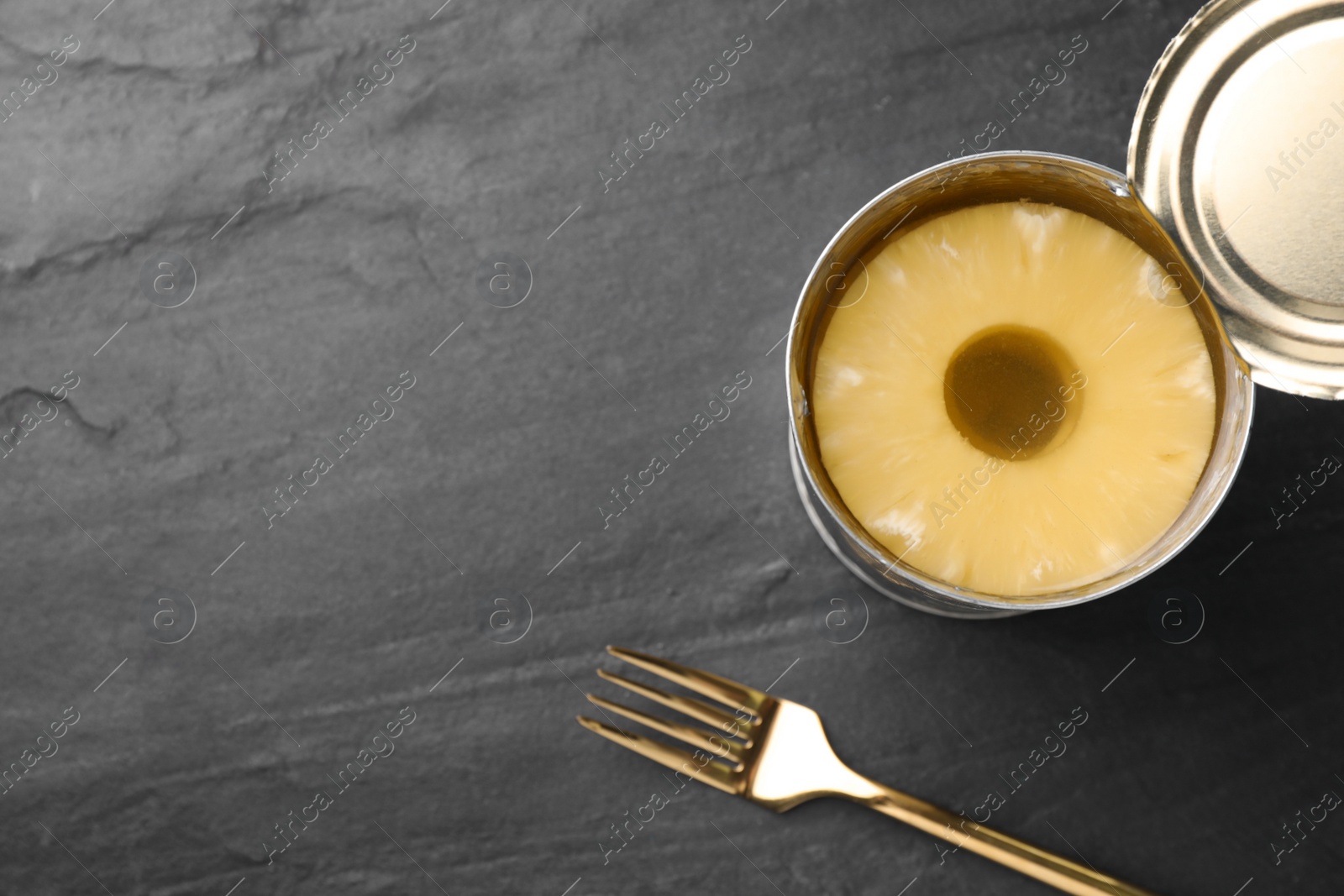 Photo of Tin with canned pineapple and fork on black slate table, flat lay. Space for text