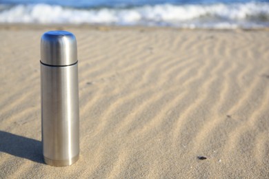 Photo of Metallic thermos with hot drink on sand near sea, space for text