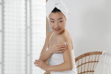 Beautiful young Asian woman applying body cream on shoulder in room