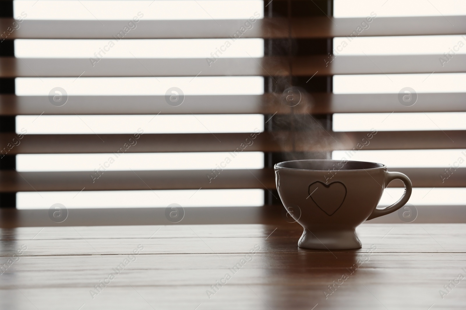 Photo of Cup of hot tea on wooden table near window with blinds. Space for text