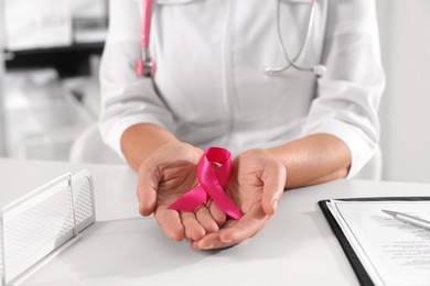Doctor holding pink ribbon at white desk indoors, closeup. Breast cancer awareness