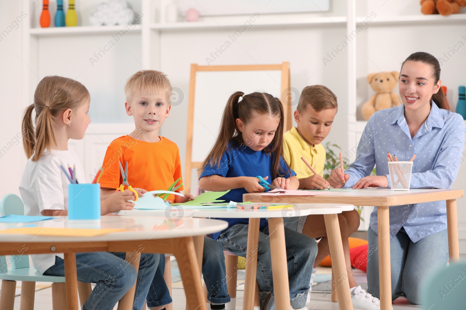 Photo of Nursery teacher with group of cute little children drawing and cutting paper at desks in kindergarten. Playtime activities