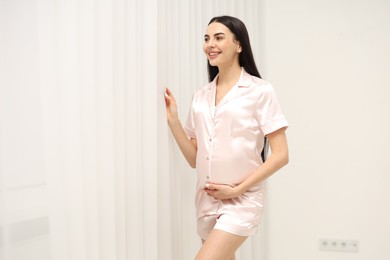 Happy pregnant woman in pajamas near window indoors