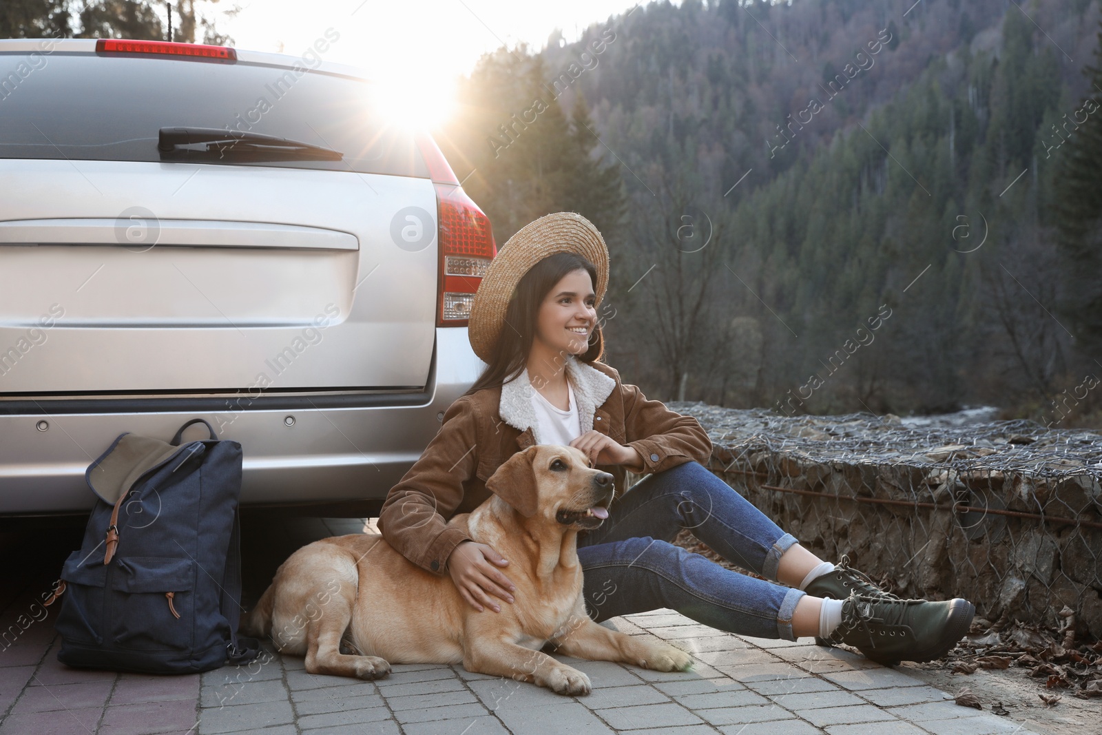 Photo of Happy woman and adorable dog sitting near car in mountains. Traveling with pet