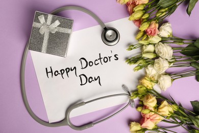 Card with phrase Happy Doctor's Day, stethoscope, gift box and flowers on purple background, flat lay