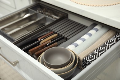 Photo of Open drawer of kitchen cabinet with different utensils, dishware and towels, closeup