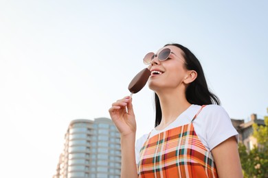 Photo of Beautiful young woman eating ice cream glazed in chocolate on city street, low angle view. Space for text