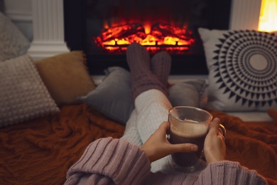 Photo of Woman with cup of coffee sitting near burning fireplace, closeup