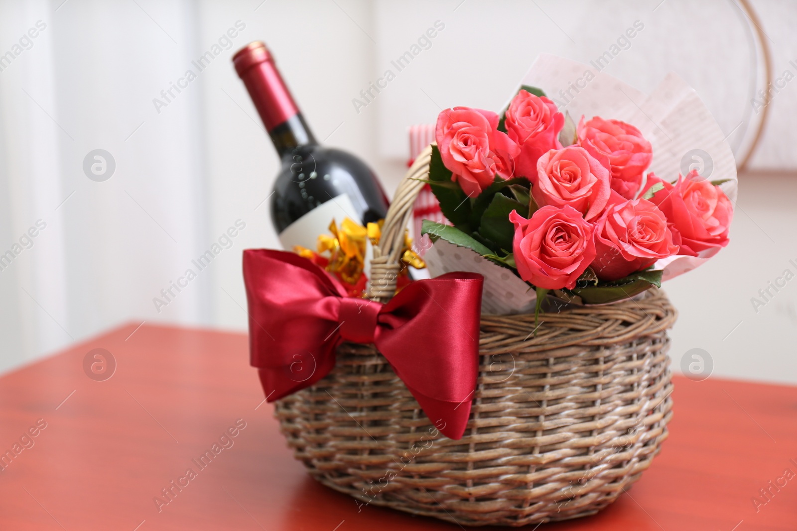 Photo of Wicker basket with gift, bouquet and wine on red table indoors