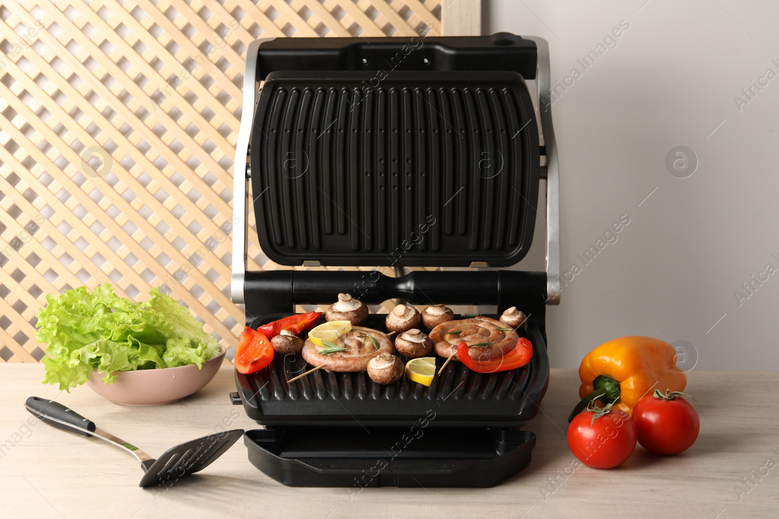 Photo of Electric grill with homemade sausages, vegetables and spatula on wooden table