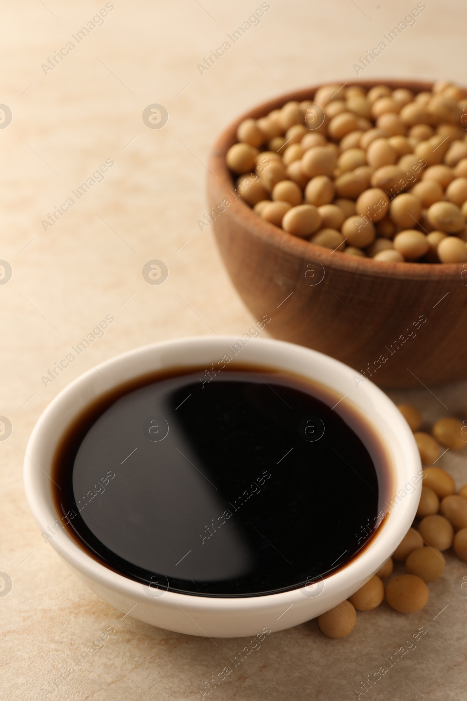 Photo of Soy sauce in bowl and soybeans on beige table, closeup