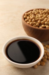 Soy sauce in bowl and soybeans on beige table, closeup