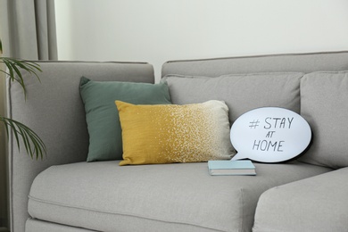 Photo of Book and speech bubble with hashtag STAY AT HOME on sofa indoors. Message to promote self-isolation during COVID‑19 pandemic