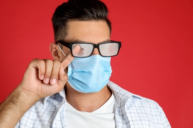 Photo of Man wiping foggy glasses caused by wearing medical mask on red background