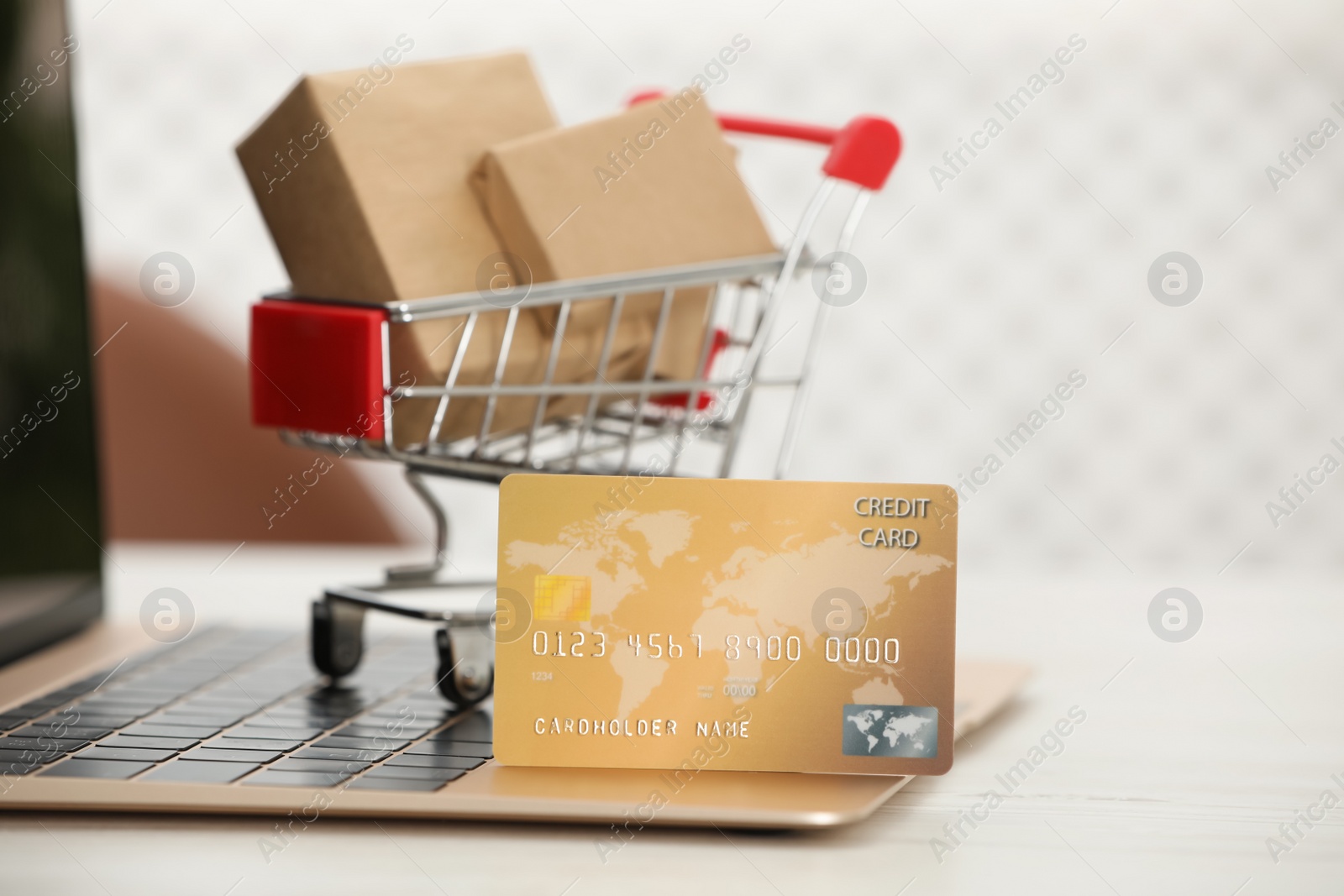 Photo of Online payment concept. Small shopping cart with bank card, boxes and laptop on white table, closeup