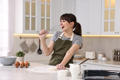 Photo of Happy young housewife with whisk having fun while cooking at white marble table in kitchen