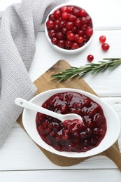 Tasty cranberry sauce in bowl, fresh berries and rosemary on white wooden table, flat lay