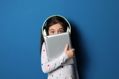 Photo of Cute little girl with headphones and tablet listening to audiobook on blue background