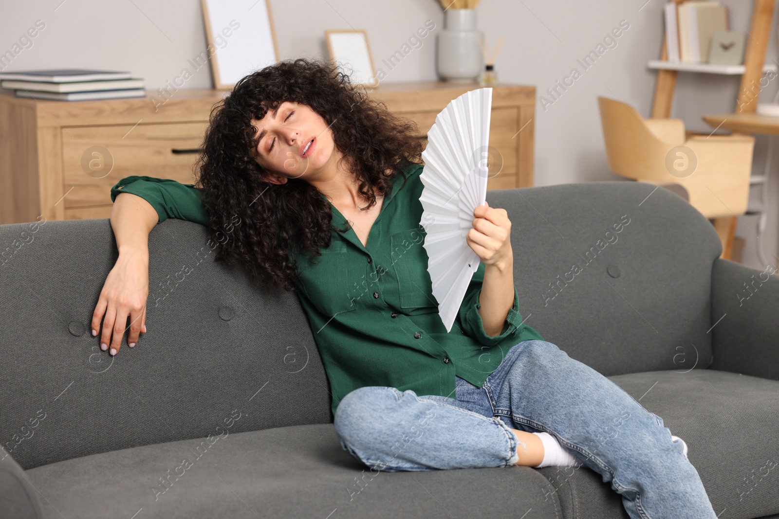 Photo of Young woman waving hand fan to cool herself on sofa at home