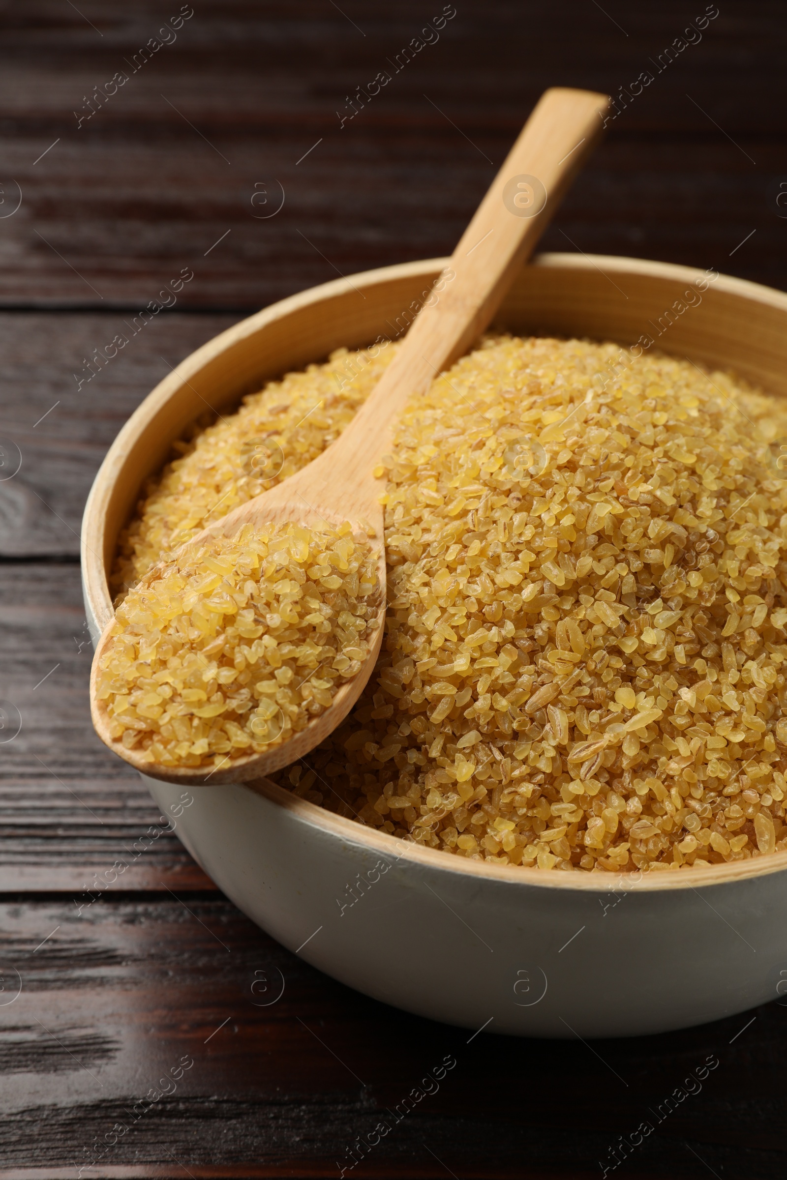 Photo of Bowl and spoon with raw bulgur on wooden table, closeup