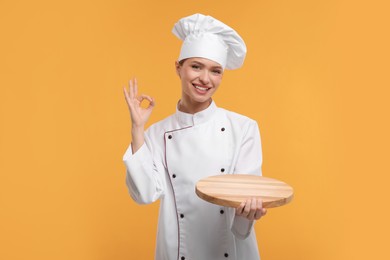 Happy chef in uniform holding empty wooden board and showing OK gesture on orange background