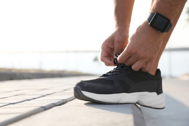 Man with fitness tracker tying shoelaces outdoors, closeup. Space for text