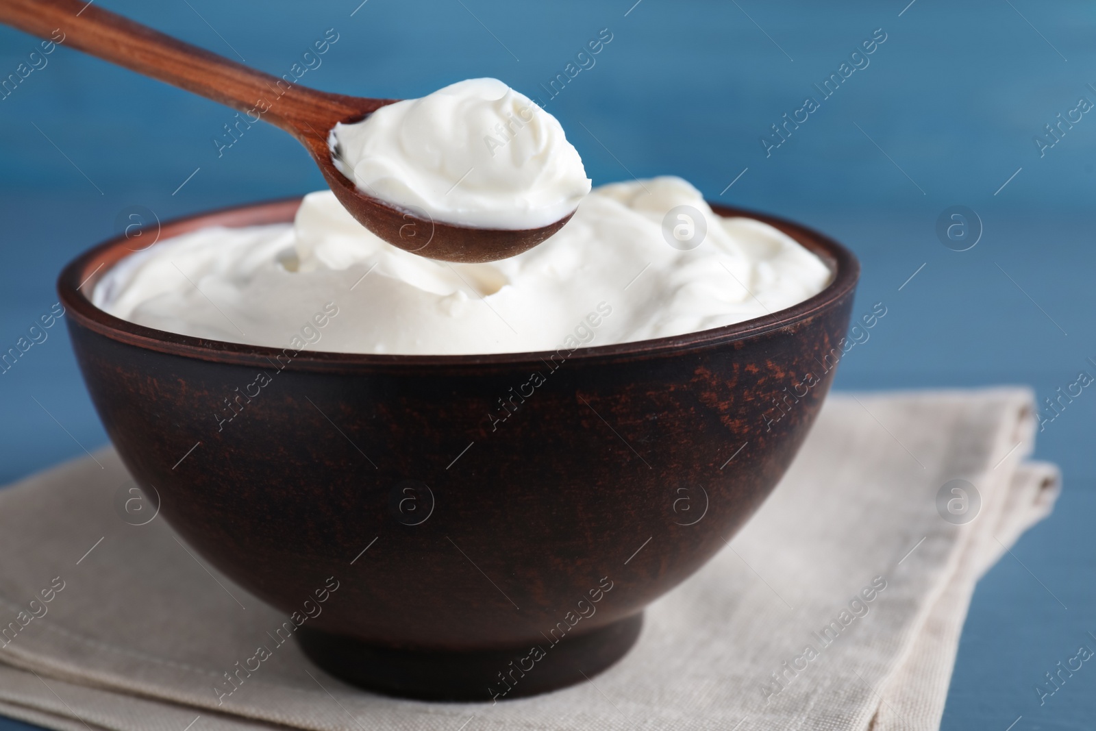 Photo of Clay spoon with sour cream above bowl on light blue wooden table, closeup