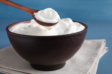 Clay spoon with sour cream above bowl on light blue wooden table, closeup