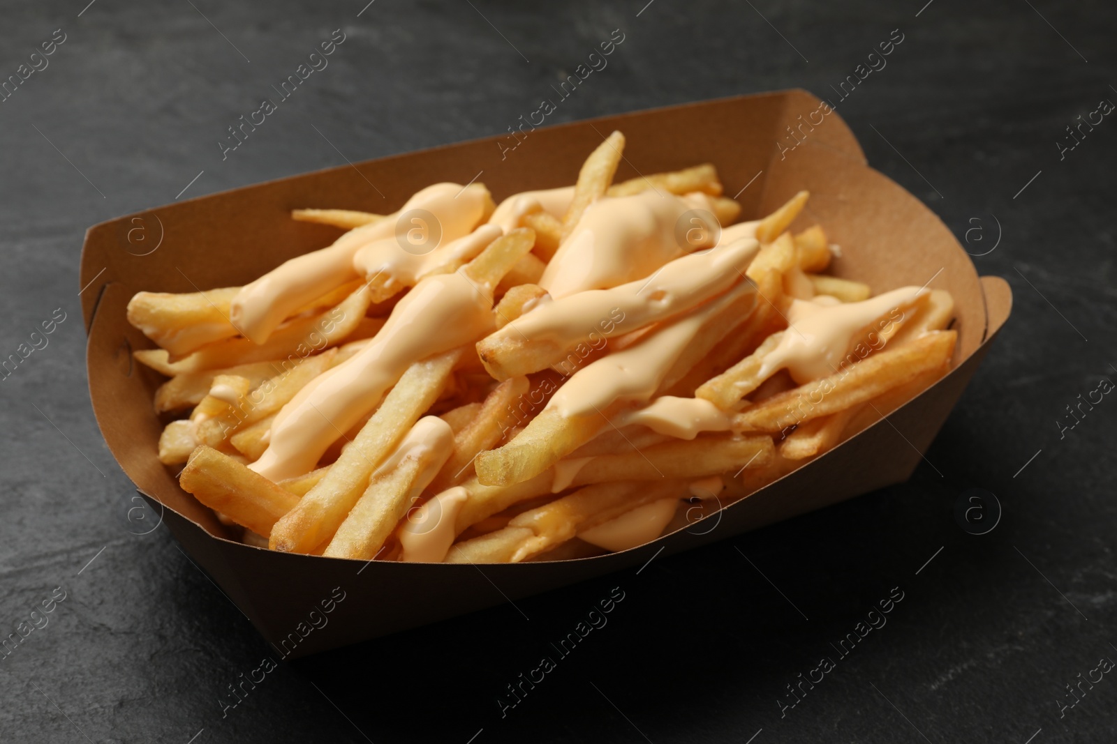 Photo of Tasty potato fries and cheese sauce in paper container on black table, closeup