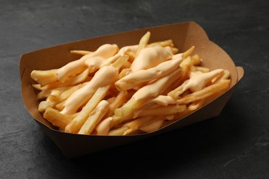 Photo of Tasty potato fries and cheese sauce in paper container on black table, closeup