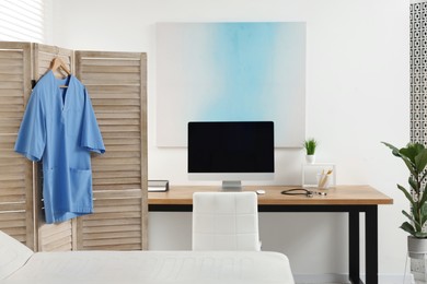 Photo of Doctor's gown hanging on wooden folding screen near workplace in clinic