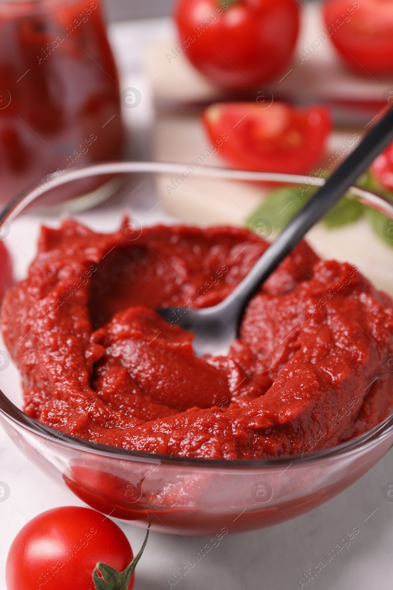 Photo of Glass bowl of tasty tomato paste with spoon and ingredients on white marble table, closeup